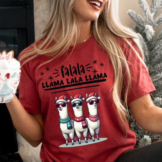 Happy, smiling woman sitting in front of a chistmas tree holding a holiday mug with marshmallows in it wearing the falala llama christmas sweatshirt in heather red featuring three llamas wearing scarfs, sunglasses, and winter hats