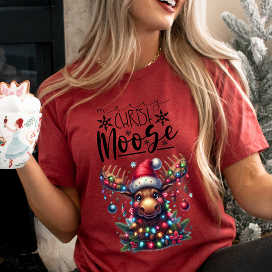 A happy, smiling woman sitting in front of a christmas tree holding a coffee mug topped with marshmallows wearing the heather red Merry Christ-moose tshirt featuring a moose decorated with ornaments and lights.