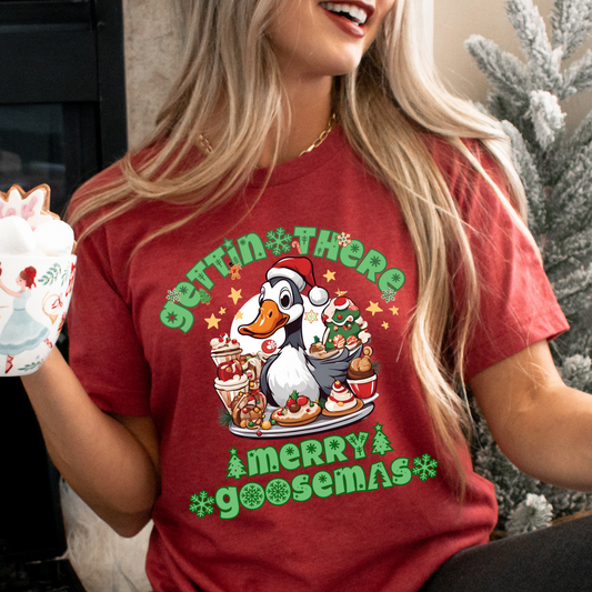 Happy, smiling woman sitting in front of christmas tree holding a holiday mug wearing the gettin there merry goosemas christmas tshirt in heather red featuring a goose with a red hat with a tray of desserts in front