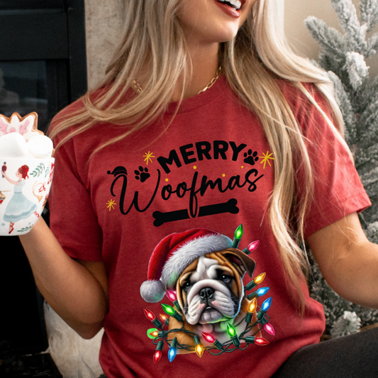 Happy, smiling woman holding a christmas mug with marshmallows on top and sitting in front of a christmas tree wearing the Merry Woofmas Christmas T-shirt in heather red featuring the words merry woofmas in black with a graphic of a dog bone below it and a boxer dog with a red hat wrapped in christmas lights