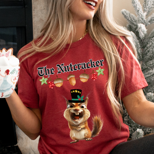 Happy, smiling woman sitting in front of a christmas tree holding a holiday mug with marshmallows on top wearing The Nutcracker christmas t-shirt in heather red featuring a funny squirrel with a black top hat with green ribbon and gold stars and a rope of acorns above