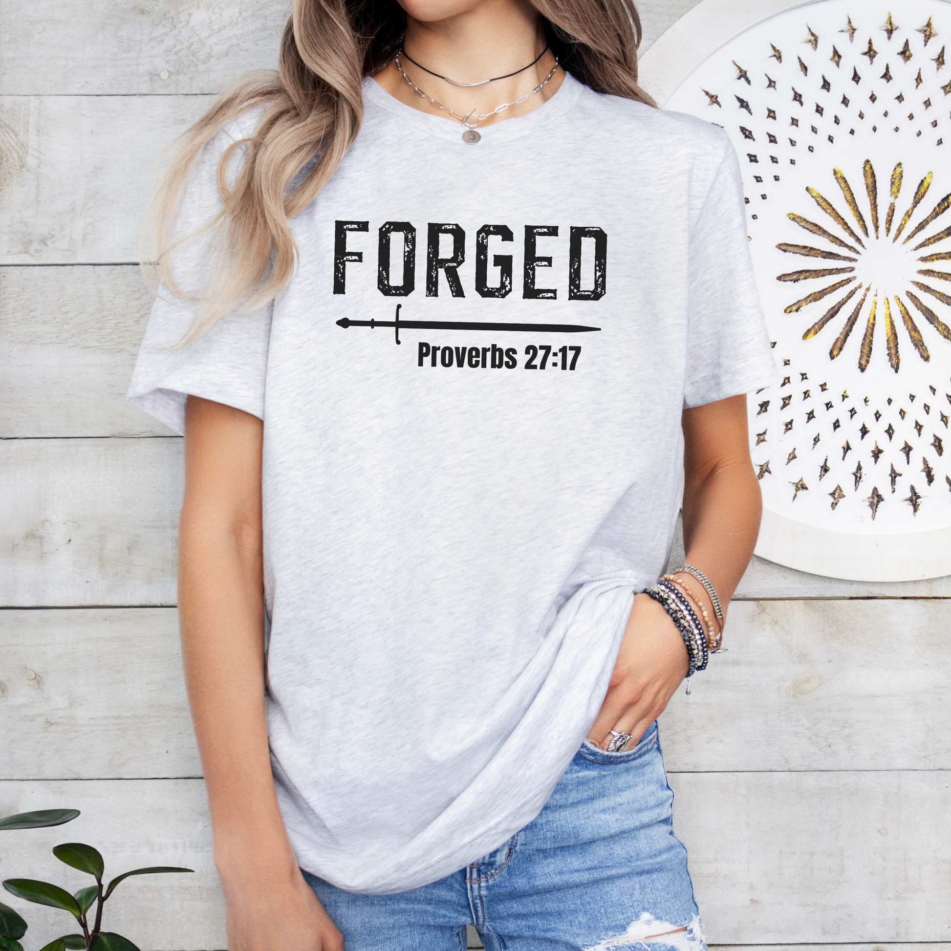 A woman standing in front of a shiplap white wall with white decor wearing the FORGE Proverbs 27:17 t-shirt in ash featuring a sword under the word FORGE and under the sword is printed Proverbs. All print is in black.