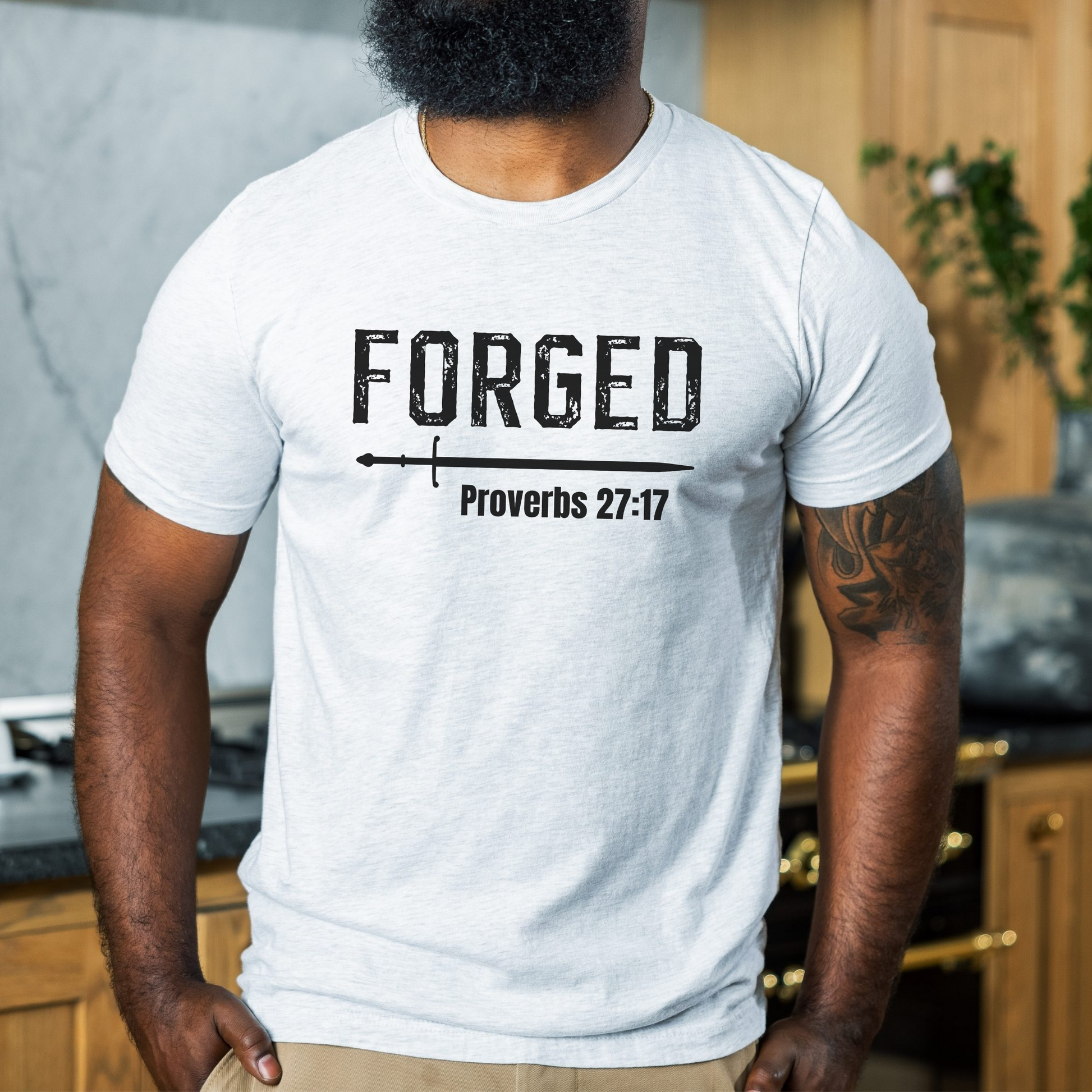 A man standing in a kitchen wearing the FORGE Proverbs 27:17 t-shirt in ash featuring a sword under the word FORGE and under the sword is printed Proverbs. All print is in black.