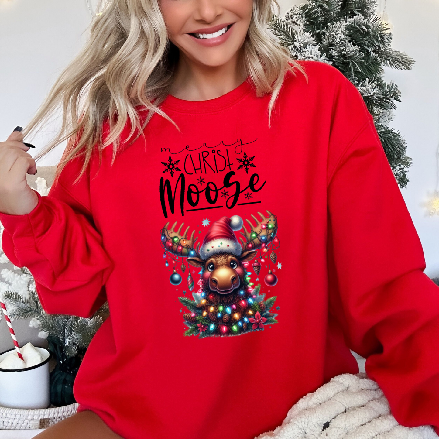 Happy, smiling woman sitting in front of a christmas tree wearing the red merry Christ moose sweatshirt in red featuring a moose decorated with christmas ornaments and lights 