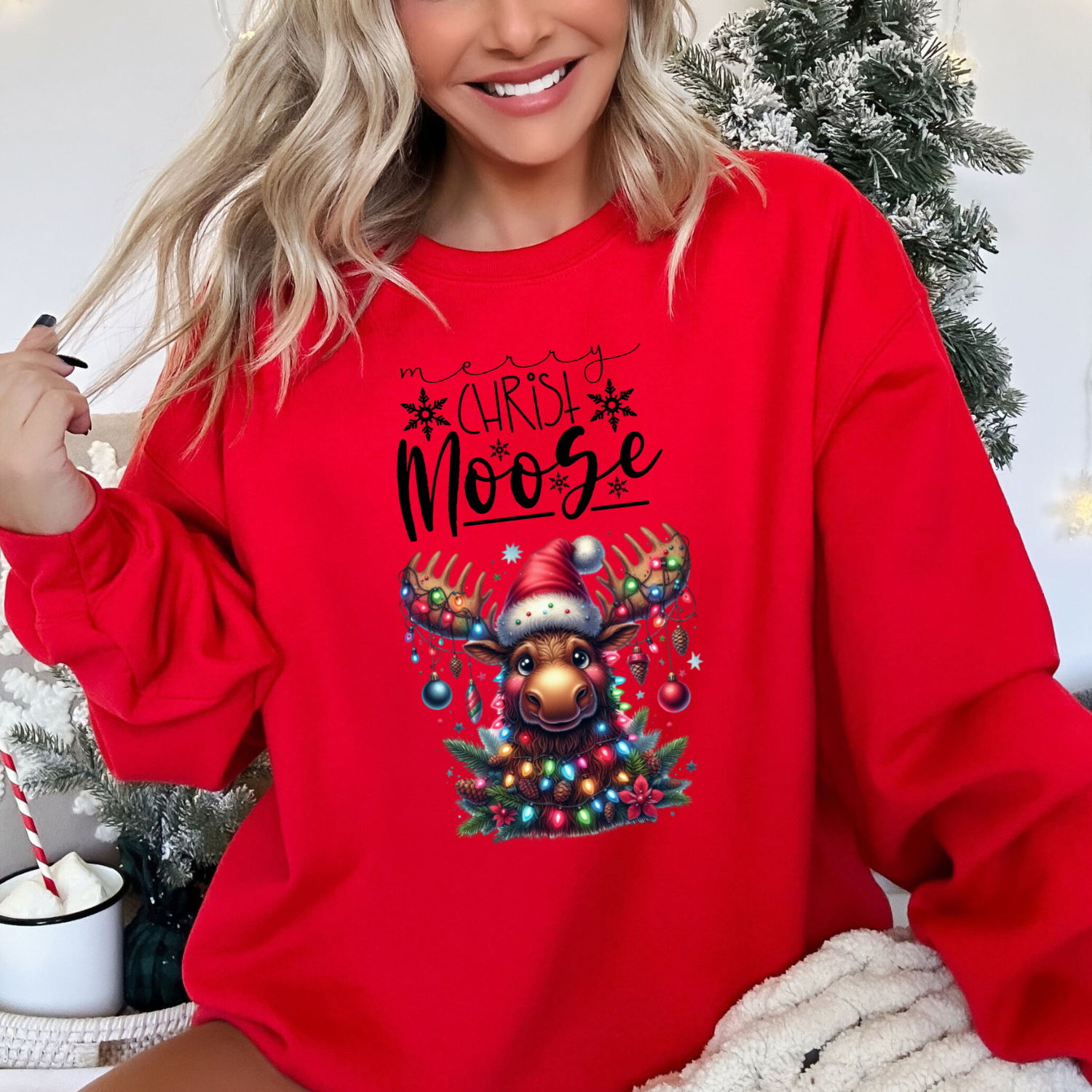 Happy, smiling woman sitting in front of a christmas tree wearing the red merry Christ moose sweatshirt in red featuring a moose decorated with christmas ornaments and lights 