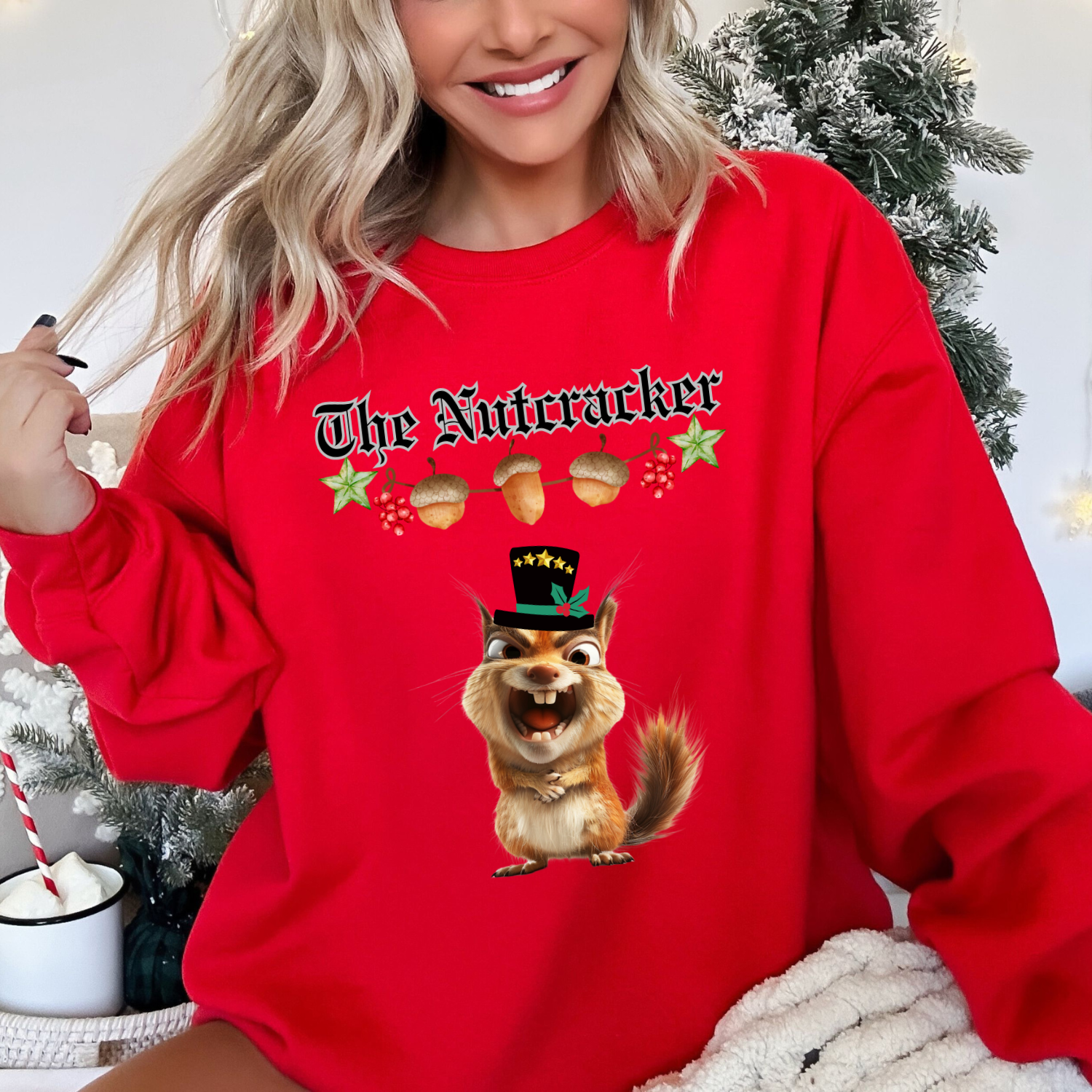 Happy, smiling woman sitting in front of a christmas tree wearing The Nutcracker christmas sweatshirt in red featuring a strand of acorns hanging above a squirrel making a goofy face wearing a black top hat with a green ribbon and gold stars