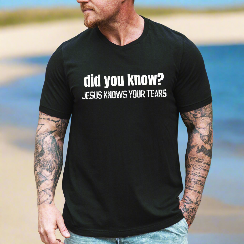 Man with tattoos on his arms standing on the beach wearing the did you know? Jesus knows your tears tshirt in black heather with text in white print