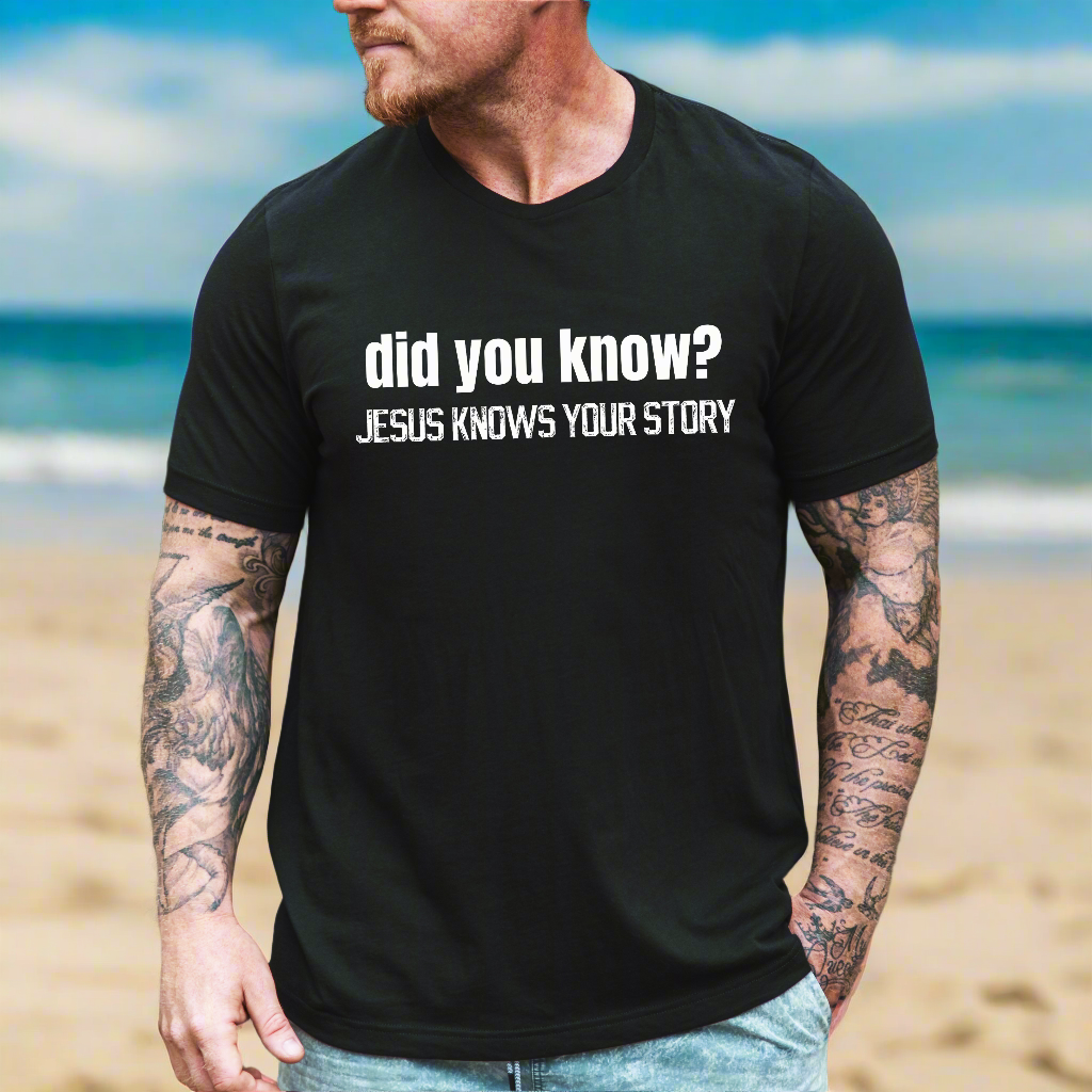 A man with tattoos on his arms standing on the beach wearing the did you know? Jesus knows your story tshirt in black heather