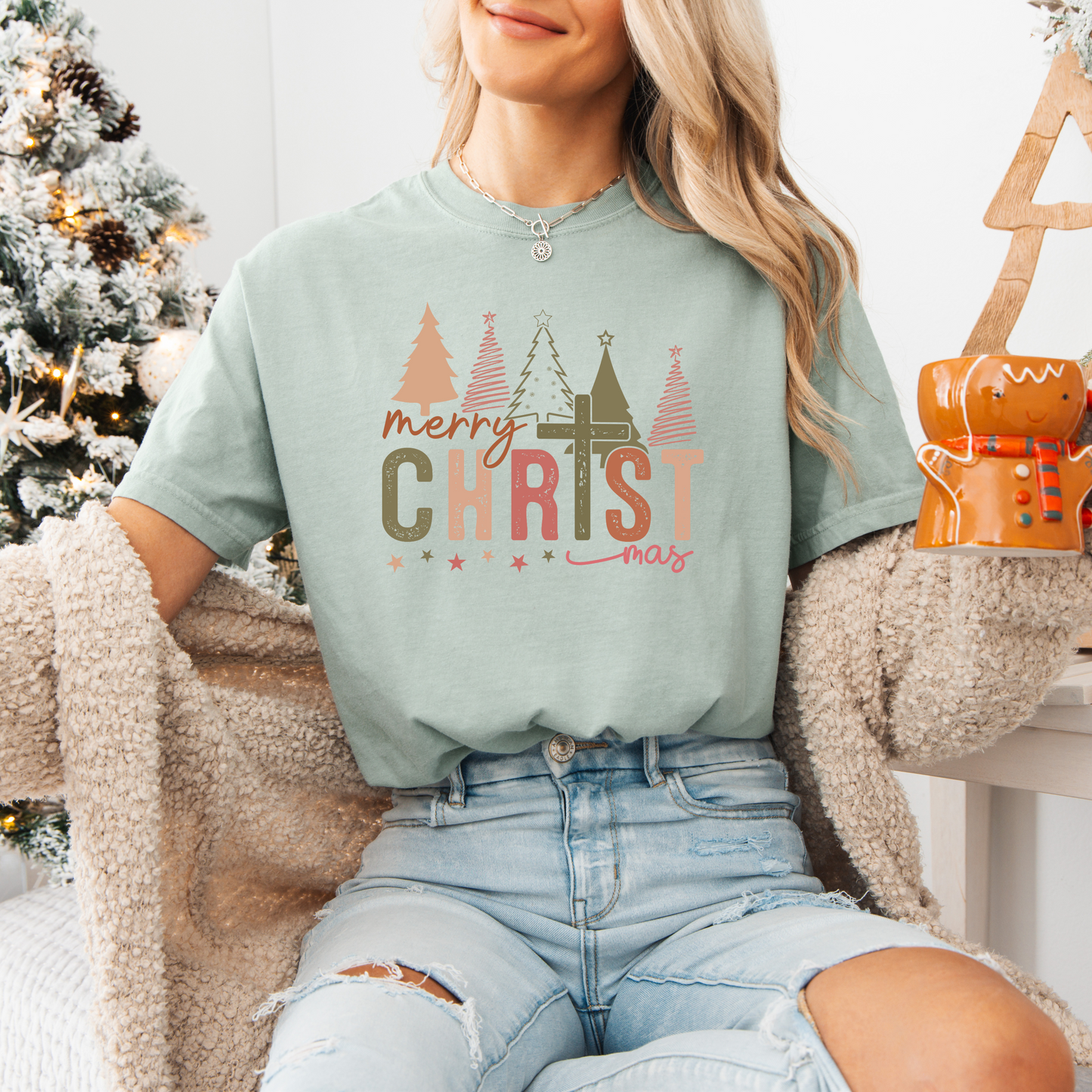 A smiling woman sitting by a christmas tree holding a gingerbread mug wearing the christmas cross shirt in bay with a cross as the T with christmas trees in the background