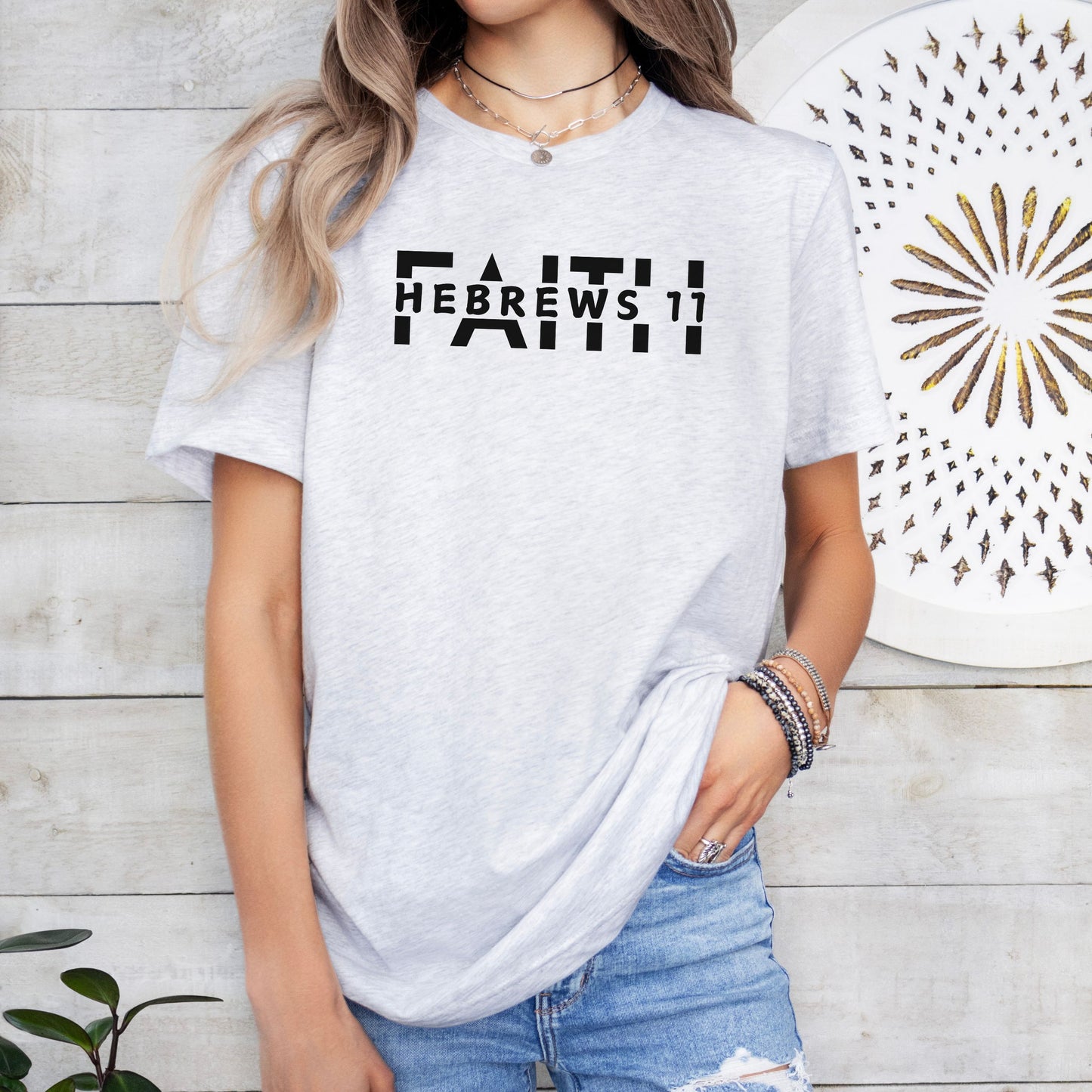 Woman standing by a fern wearing the FAITH Hebrews 11 tshirt in ash featuring the word faith written in capitals split in half with the Hebrews 11 in capitals all in black print.