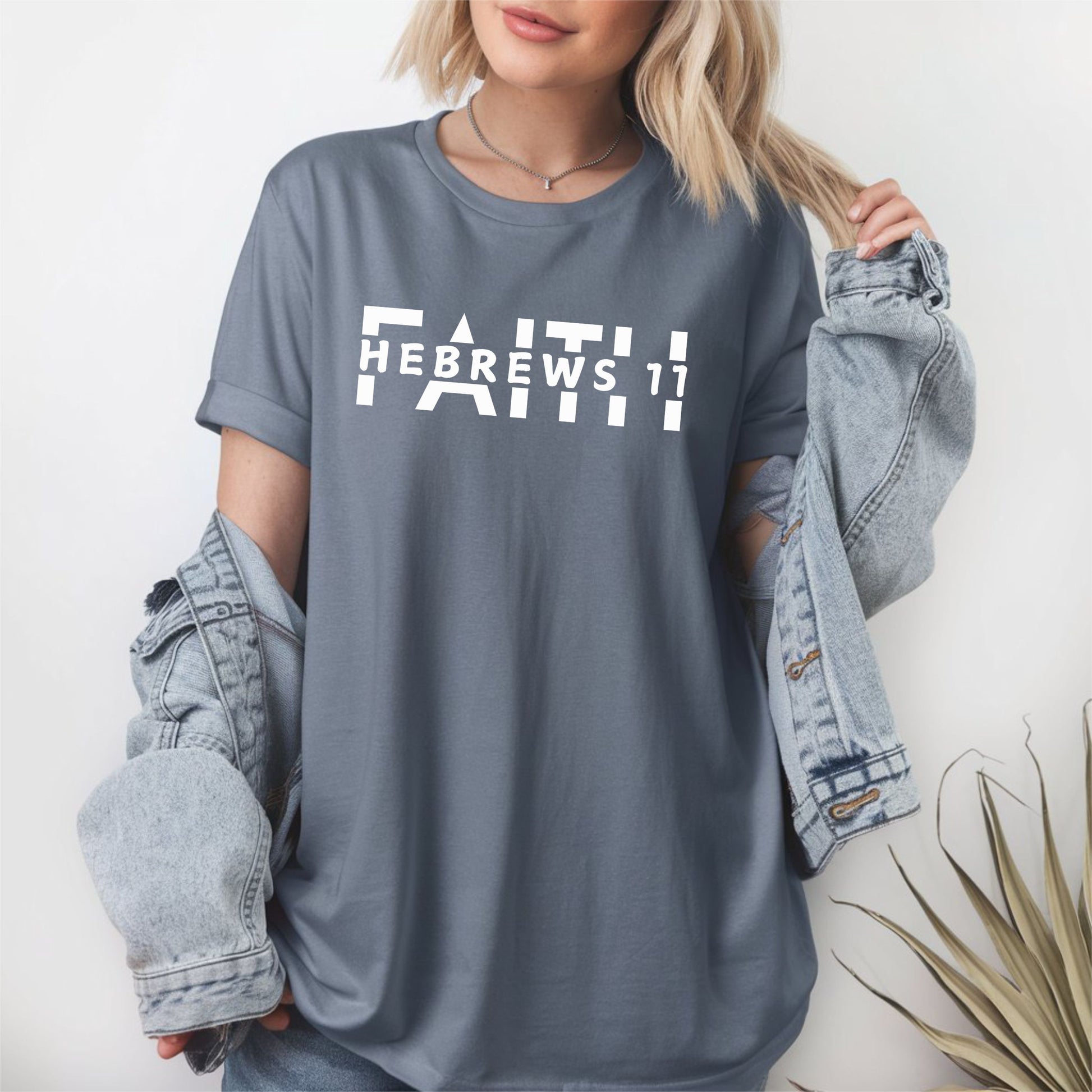 Woman standing by a fern wearing the FAITH Hebrews 11 graphite tshirt in  featuring the word faith written in capitals split in half with the Hebrews 11 in capitals all in white print.