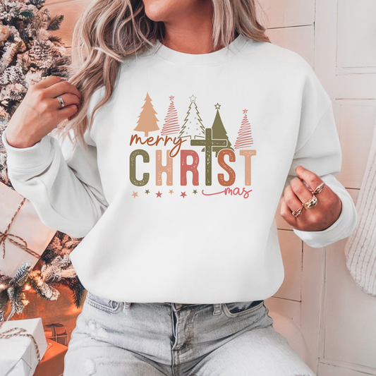 A woman sitting by a christmas tree wearing the christmas cross sweatshirt in white featuring a cross for the T and christmas trees above the wording