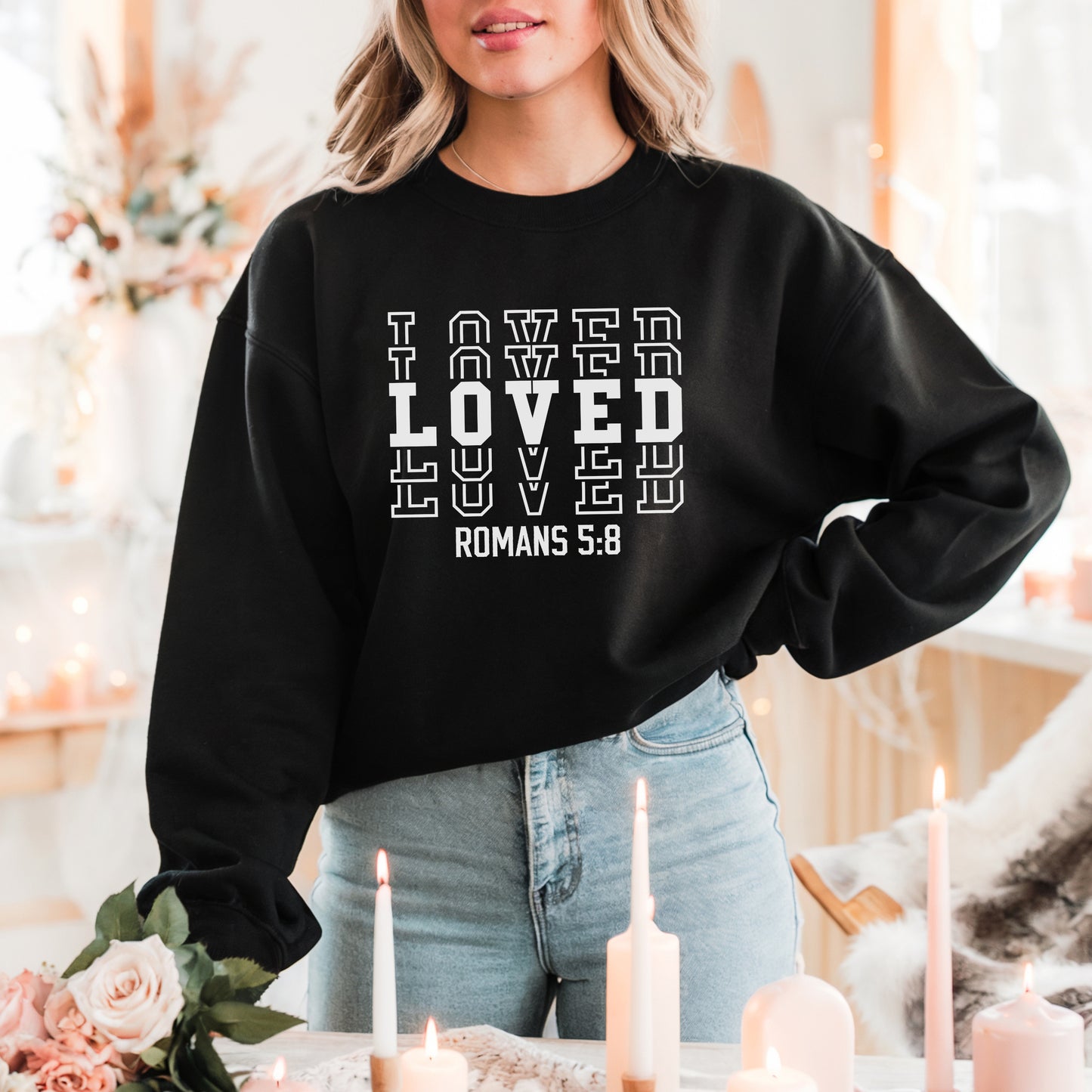 Woman standing in front of white and light pink floral decor wearing the LOVED Romans 5:8 sweatshirt in black with white print