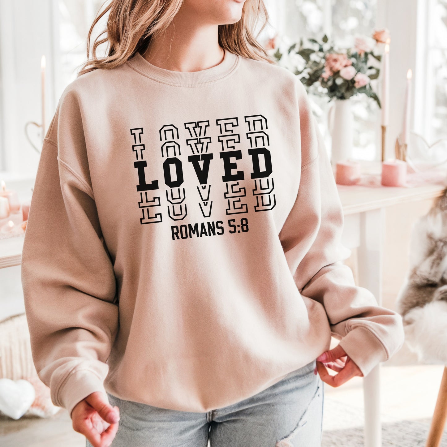 Woman standing in front of white and light pink floral decor wearing the LOVED Romans 5:8 sweatshirt in sand with black print