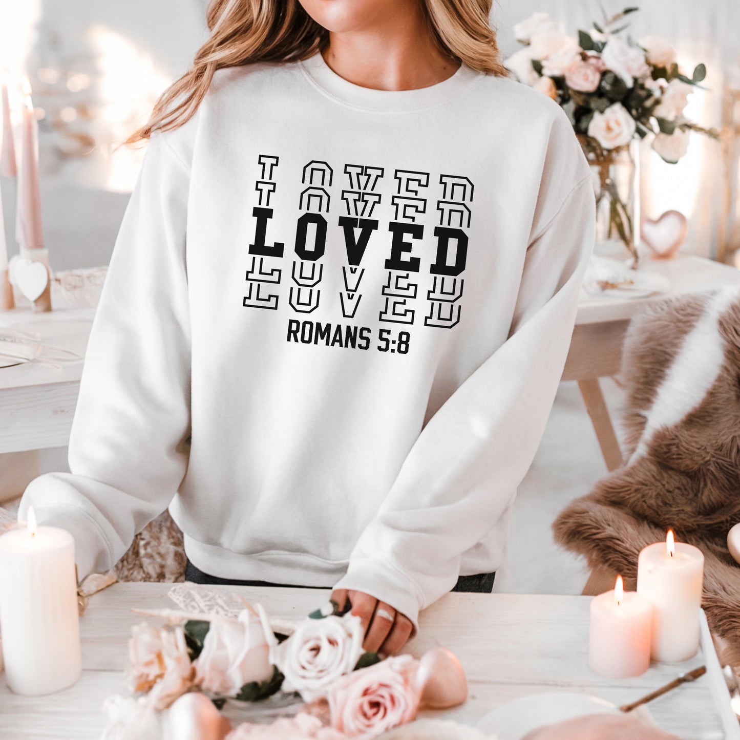 Woman standing in front of white and light pink floral decor wearing the LOVED Romans 5:8 sweatshirt in white with black print