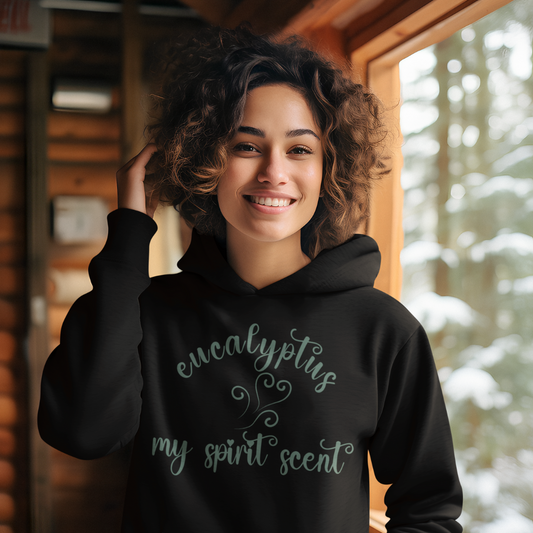 A smiling woman in a cozy winter cabin wearing the My Spirit Scent is Eucalyptus hoodie in black with script font and an aroma script heart both in a blue-green hue