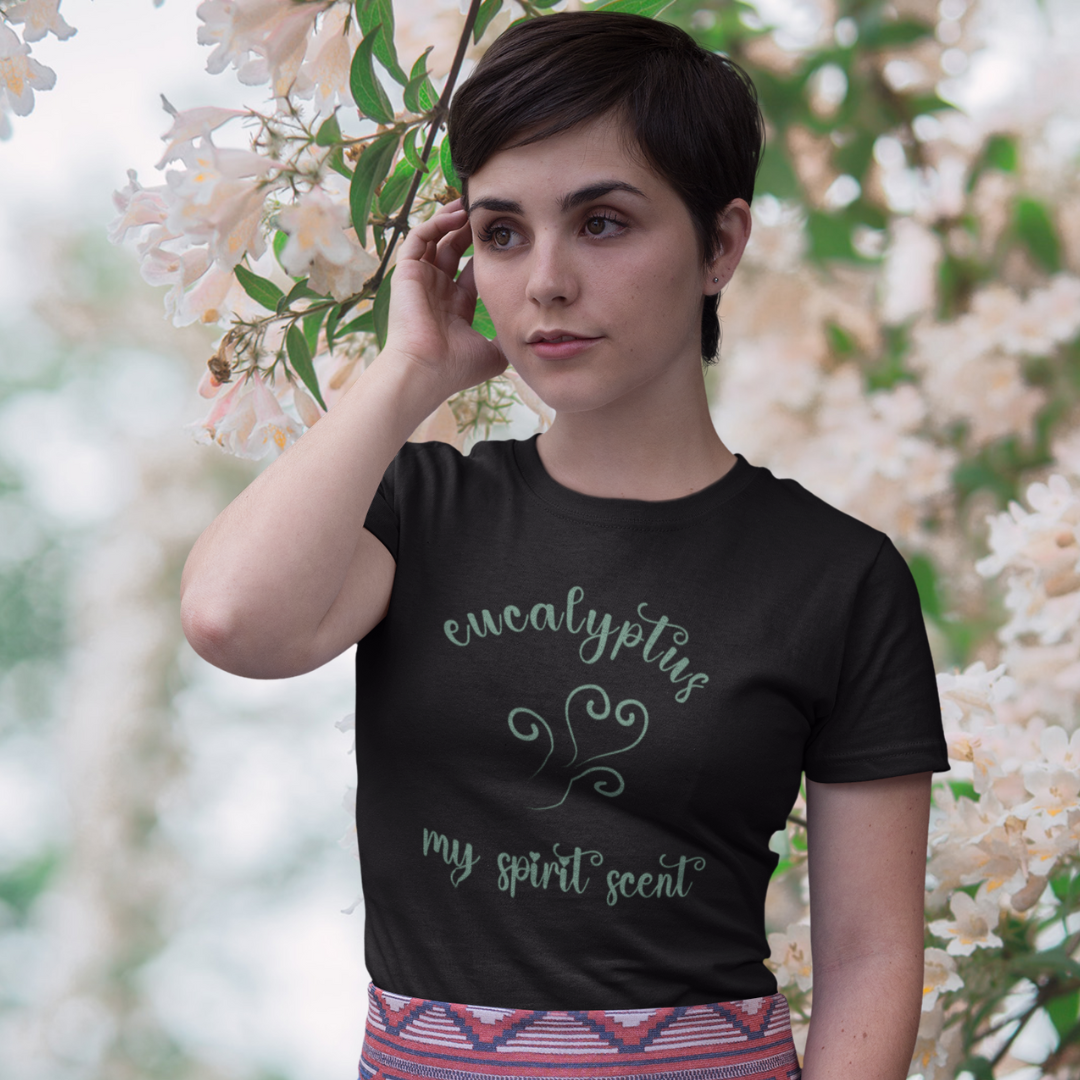 A woman standing rather curiously in front of a cherry blossom backdrop wearing the My Spirit Scent is Eucalyptus t-shirt in black heather with script font and an aroma script heart both in a blue-green hue