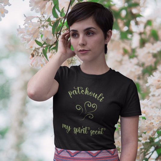 A woman standing rather curiously in front of a cherry blossom backdrop wearing the My Spirit Scent is Patchouli t-shirt in black heather with script font and an aroma script heart both in a mossy green hue