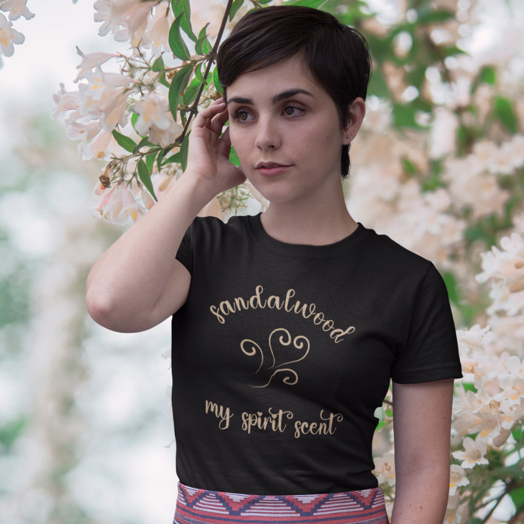 A woman standing rather curiously in front of a cherry blossom backdrop wearing the My Spirit Scent is Sandalwood t-shirt in black heather with script font and an aroma script heart both in a warm, muted beige hue