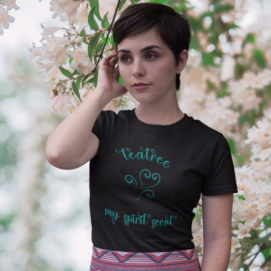 A woman standing rather curiously in front of a cherry blossom backdrop wearing the My Spirit Scent is Tea Tree t-shirt in black heather with script font and an aroma script heart both in a teal hue