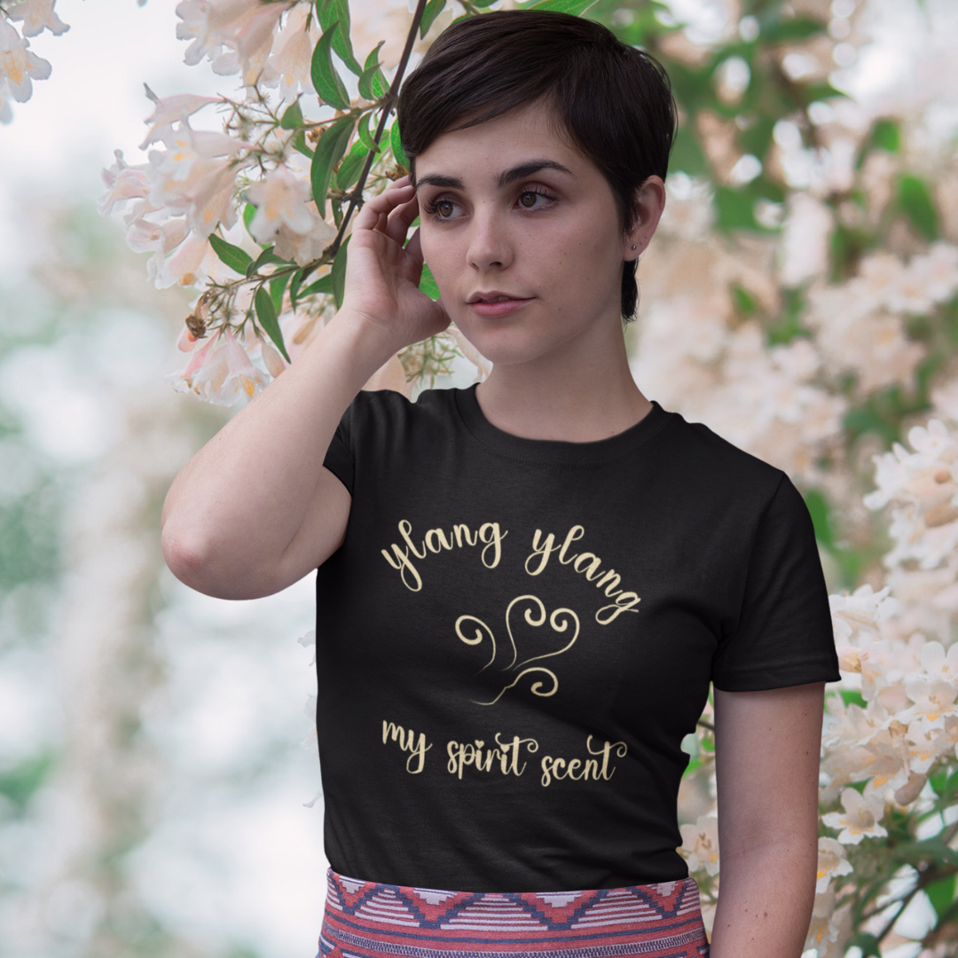 A woman standing rather curiously in front of a cherry blossom backdrop wearing the My Spirit Scent is Ylang Ylang t-shirt in black heather with script font and an aroma script heart both in a soft, pastel yellow hue