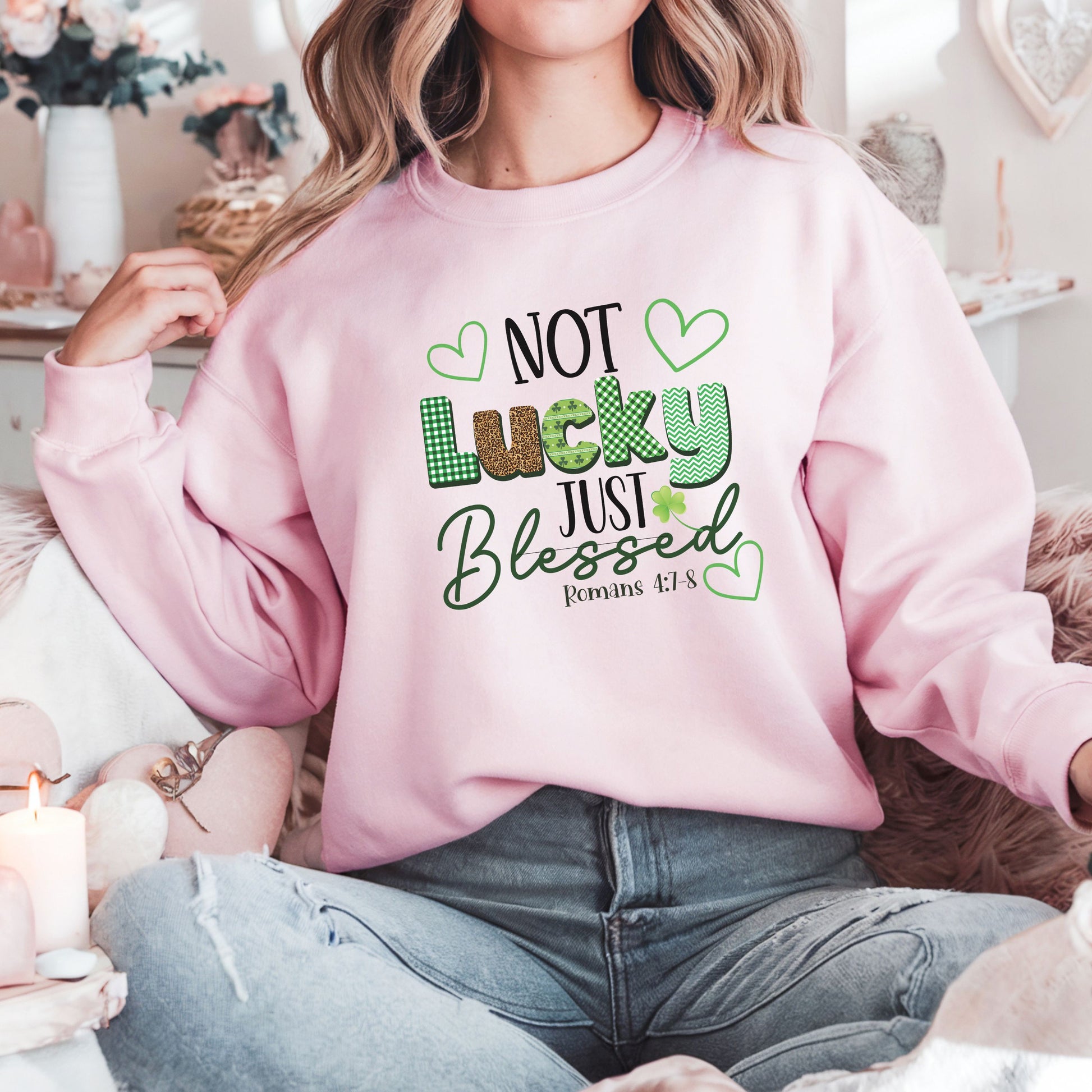 A woman sitting in front of white and pink floral decor wearing the not luck just blessed sweatshirt in light pink featuring romans 4:7-8 on it as well