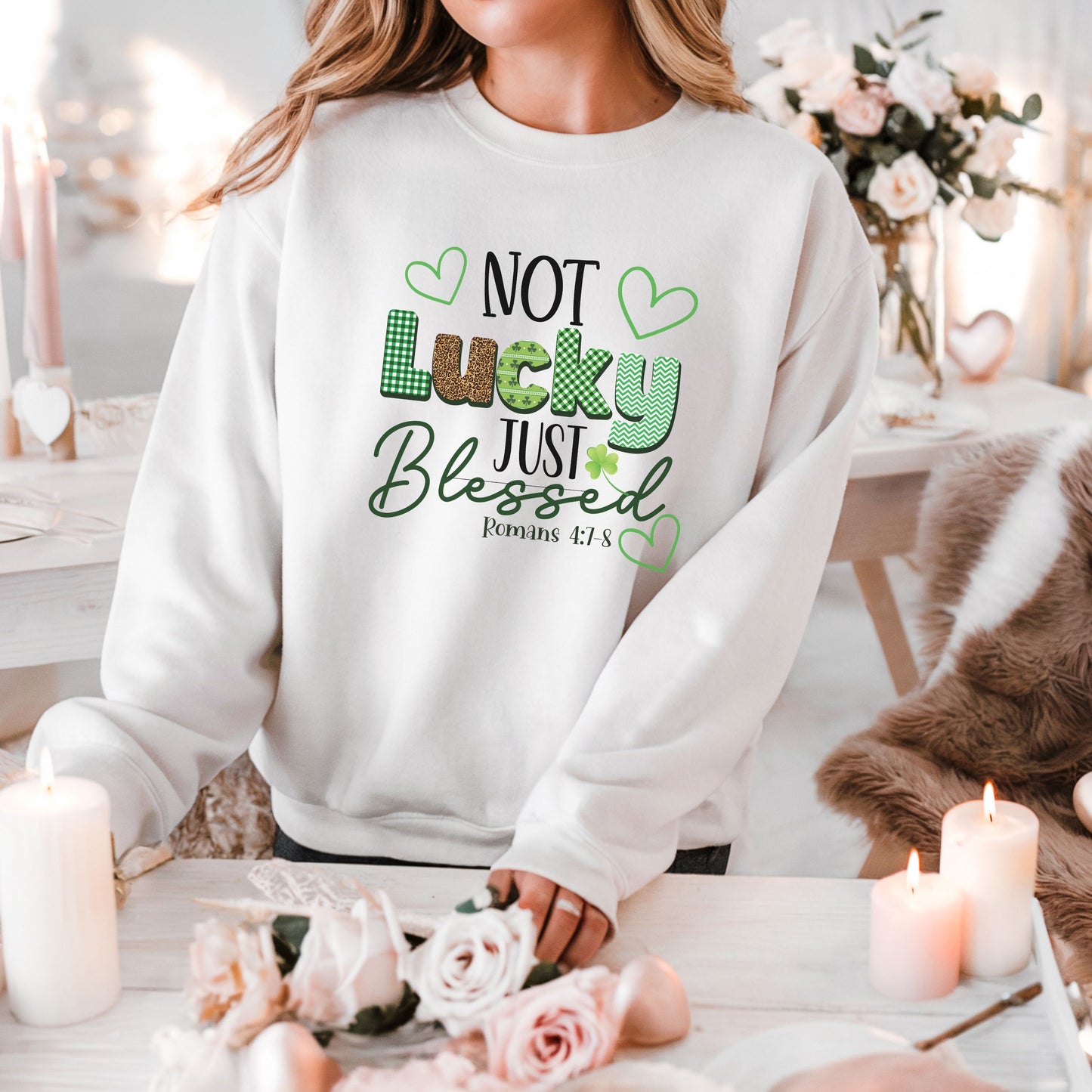 A woman standing in front of white and pink floral decor wearing the not luck just blessed sweatshirt in white featuring romans 4:7-8 on it as well