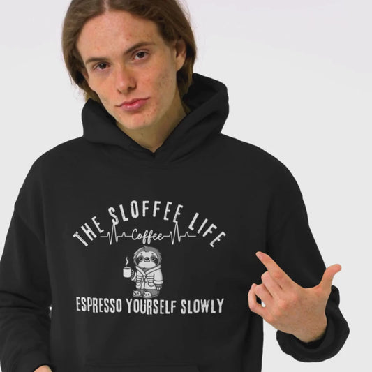 Happy young man in a studio setting wearing The Sloffee Life Sleepzz Coffeezz Repeatzz hoodie in black featuring a sloth lying on the ground holding a coffee