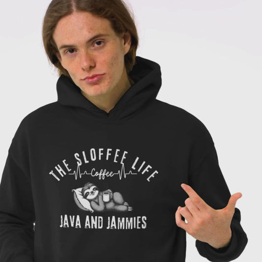 Happy young man in a studio setting wearing The Sloffee Life Java and Jammies hoodie in black featuring a sloth relaxing on a pillow holding a mug of coffee