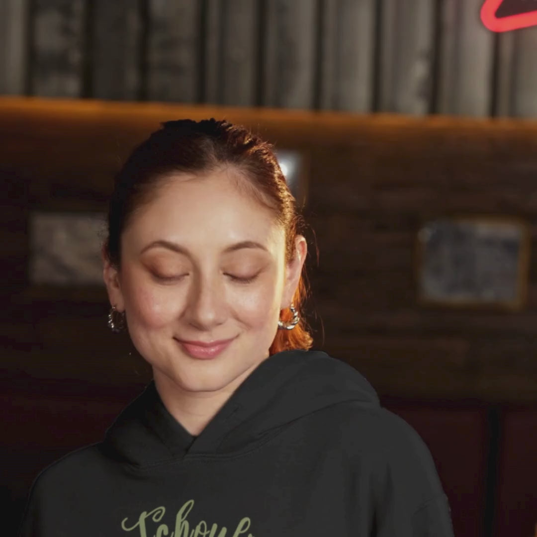 A smiling woman standing in a bar and grill wearing the My Spirit Scent is Patchouli hoodie in black with script font and an aroma script heart both in a mossy green hue