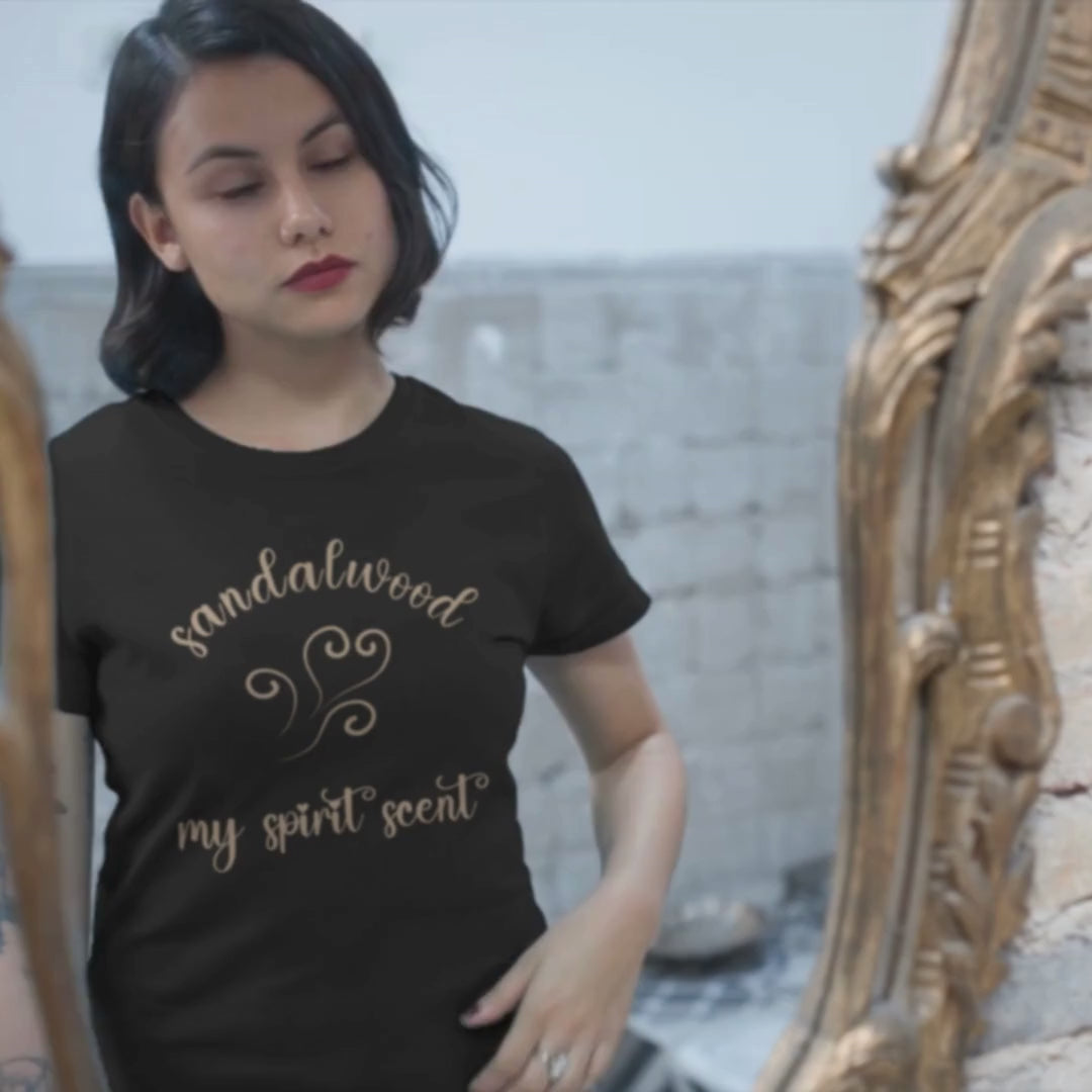 A woman looking at her reflection in an antique mirror wearing the My Spirit Scent is Sandalwood t-shirt in black heather with script font and an aroma script heart both in a warm, muted beige hue