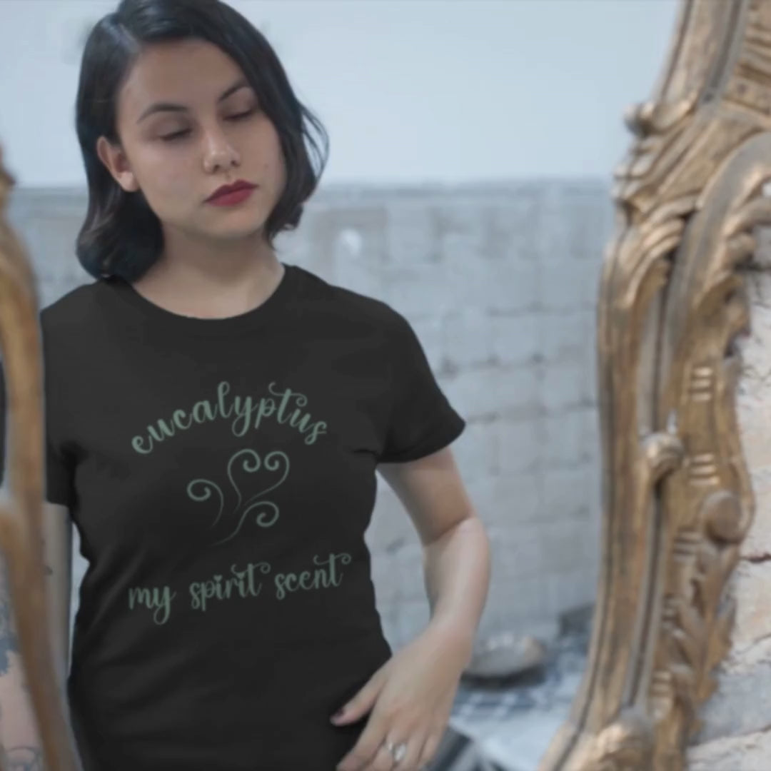 A woman looking at her reflection in an antique mirror wearing the My Spirit Scent is Eucalyptus t-shirt in black heather with script font and an aroma script heart both in a blue-green hue