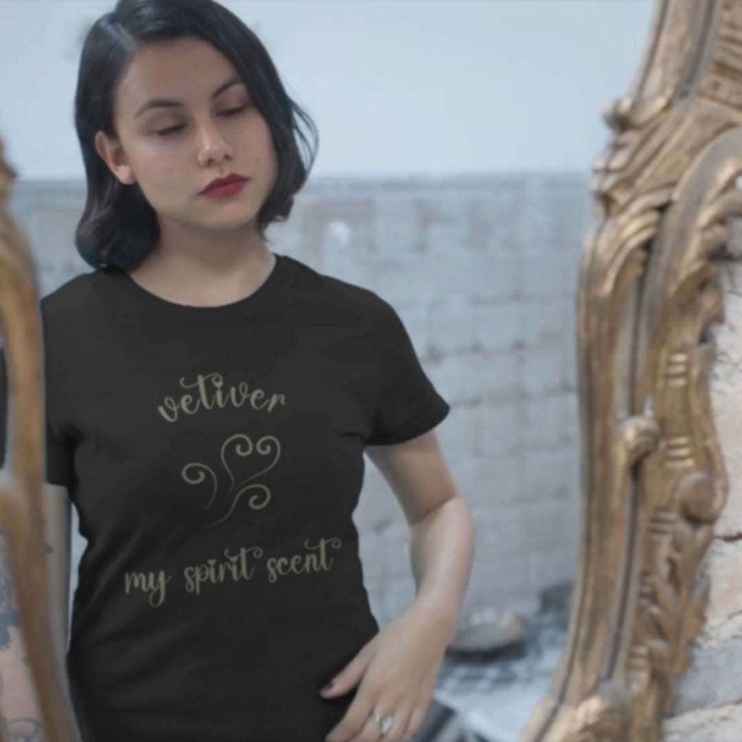 A woman looking at her reflection in an antique mirror wearing the My Spirit Scent is Vetiver t-shirt in black heather with script font and an aroma script heart both in a grey-green hue