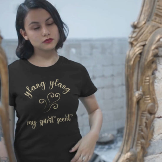 A woman looking at her reflection in an antique mirror wearing the My Spirit Scent is Ylang Ylang t-shirt in black heather with script font and an aroma script heart both in a soft, pastel yellow hue