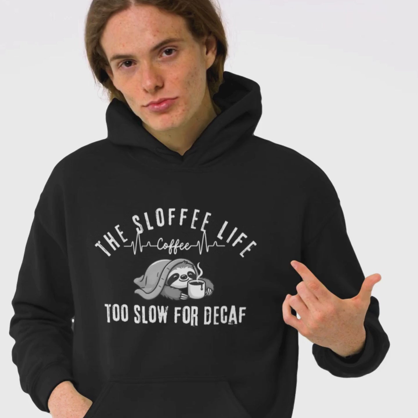 Happy young man in a studio setting wearing The Sloffee Life Too Slow For Decaf hoodie in dark heather featuring a sloth under a blanket holding a coffee mug