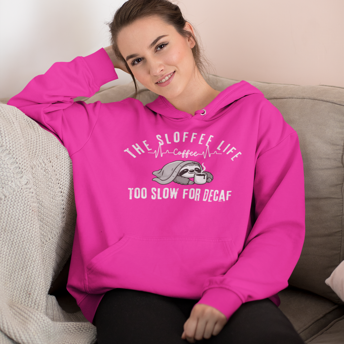 Comfortable woman relaxing at home wearing The Sloffee Life Too Slow For Decaf hoodie in heliconia featuring a sloth under a blanket holding a coffee cup